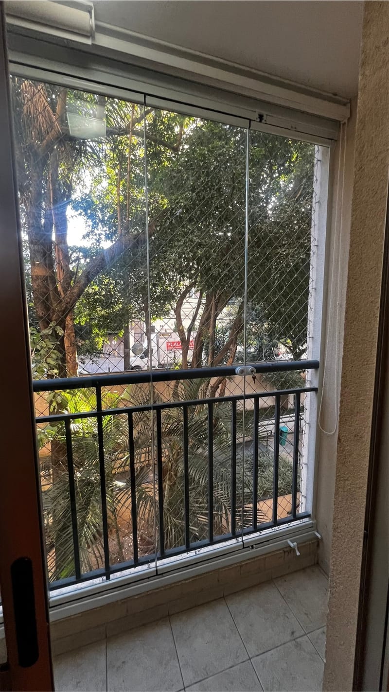 Varanda de apartamento fechada com vidro e tela de proteção, com vista para área arborizada. Obra da VitraAlu em Piqueri, São Paulo.